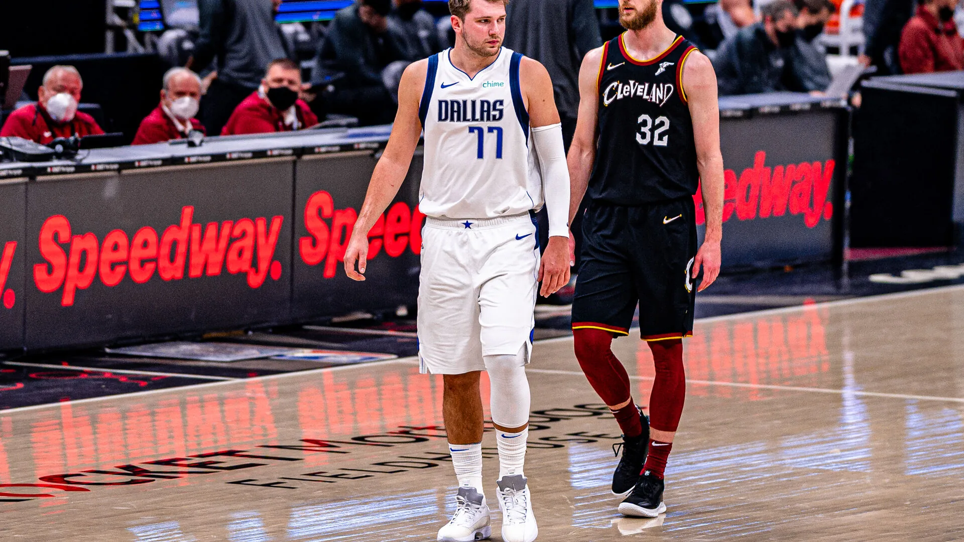 NBA game at Rocket Mortgage FieldHouse: Luka Doncic in the white Dallas Mavericks kit with a chime chest patch walking beside Dean Wade in the black Cleveland Cavaliers kit, red Speedway LED boards lining the court behind them