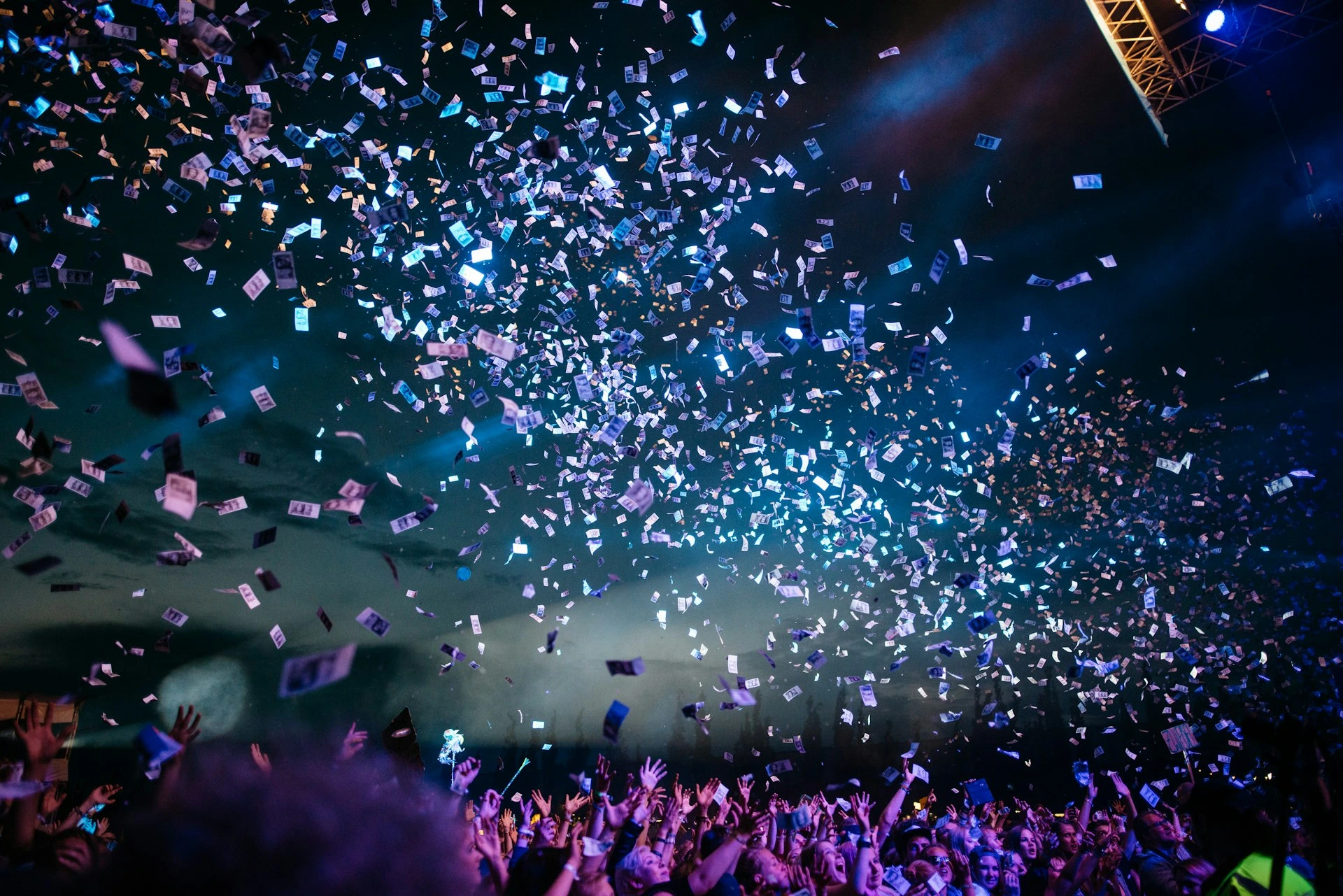 Confetti bursting over a festival crowd with raised hands under blue stage lights at night