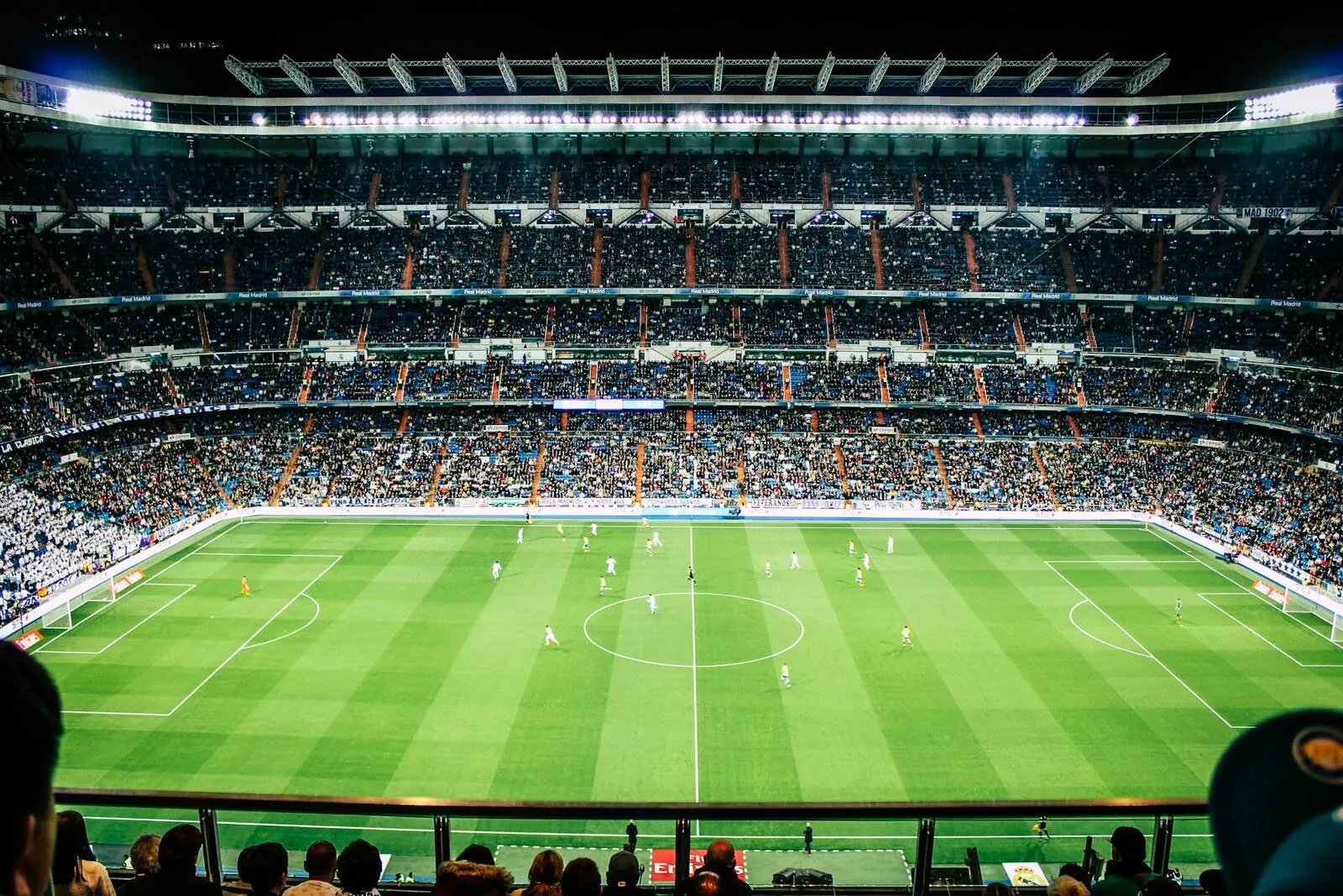 Packed football stadium at night seen from the stands, sponsor boards circling the floodlit pitch